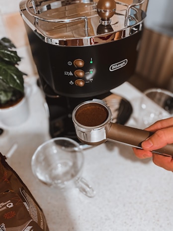 A hand holds a portafilter filled with ground coffee in front of a DeLonghi espresso machine. The machine is black with metallic accents and has several buttons, a cup icon, and a glowing light. Nearby, there is a clear glass cup and a package of coffee beans. A potted plant is partially visible on the side.