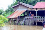 A wooden stilt house with a metal roof is surrounded by floodwaters. Two people stand on the porch, looking out at the water. Lush green foliage and banana plants are visible in the background.