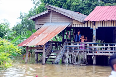 A wooden stilt house with a metal roof is surrounded by floodwaters. Two people stand on the porch, looking out at the water. Lush green foliage and banana plants are visible in the background.