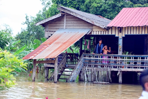 A wooden stilt house with a metal roof is surrounded by floodwaters. Two people stand on the porch, looking out at the water. Lush green foliage and banana plants are visible in the background.
