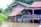 A wooden stilt house with a metal roof is surrounded by floodwaters. Two people stand on the porch, looking out at the water. Lush green foliage and banana plants are visible in the background.
