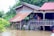 A wooden stilt house with a metal roof is surrounded by floodwaters. Two people stand on the porch, looking out at the water. Lush green foliage and banana plants are visible in the background.