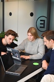 Three people are gathered around a table, each with a laptop open in front of them. They appear focused and engaged in a discussion or collaborative work. One person is pointing at the screen, possibly explaining or demonstrating something to the others. The room has modern decor with a frosted glass wall featuring minimalistic design elements.