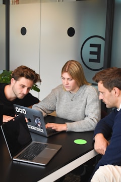 Three people are gathered around a table, each with a laptop open in front of them. They appear focused and engaged in a discussion or collaborative work. One person is pointing at the screen, possibly explaining or demonstrating something to the others. The room has modern decor with a frosted glass wall featuring minimalistic design elements.