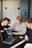 Three people are gathered around a table, each with a laptop open in front of them. They appear focused and engaged in a discussion or collaborative work. One person is pointing at the screen, possibly explaining or demonstrating something to the others. The room has modern decor with a frosted glass wall featuring minimalistic design elements.