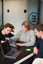 Three people are gathered around a table, each with a laptop open in front of them. They appear focused and engaged in a discussion or collaborative work. One person is pointing at the screen, possibly explaining or demonstrating something to the others. The room has modern decor with a frosted glass wall featuring minimalistic design elements.