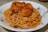 A plate filled with spaghetti coated in a rich tomato-based sauce mixed with chunks of seasoned chicken. The dish is served in a white bowl featuring floral patterns along the rim.