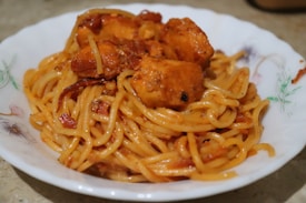 A plate filled with spaghetti coated in a rich tomato-based sauce mixed with chunks of seasoned chicken. The dish is served in a white bowl featuring floral patterns along the rim.