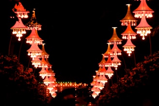 A series of ornate, vivid pink and orange lit lanterns hang in a symmetrical arrangement against a dark background. These lanterns create a luminous pathway, illuminating the surroundings with a warm glow.