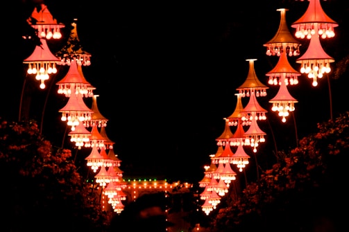 A series of ornate, vivid pink and orange lit lanterns hang in a symmetrical arrangement against a dark background. These lanterns create a luminous pathway, illuminating the surroundings with a warm glow.