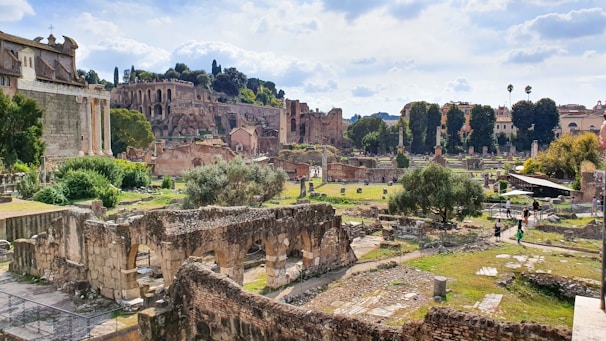 Ancient ruins and archaeological structures are surrounded by lush greenery and trees, with a scenic backdrop of hills and historical buildings under a partly cloudy sky. Pathways and scattered stones enhance the sense of historical significance.
