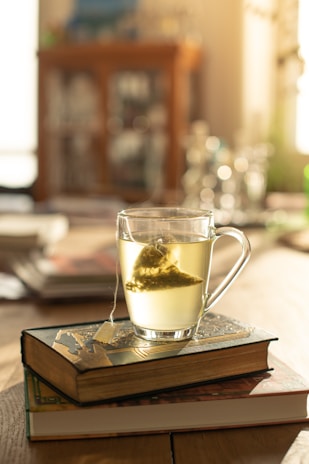A glass cup filled with light-colored tea is placed on top of two closed, ornate books. The teabag string drapes over the side of the cup. The setting appears to be a warm, sunlit room, with a blurred background suggesting shelves or furniture.