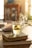 A glass cup filled with light-colored tea is placed on top of two closed, ornate books. The teabag string drapes over the side of the cup. The setting appears to be a warm, sunlit room, with a blurred background suggesting shelves or furniture.
