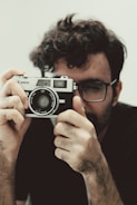 A person is holding a vintage Canon film camera up to their face, peering through the viewfinder. The individual has curly dark hair and is wearing glasses. The camera is prominently displayed, creating a sense of focus on photography and vintage technology.