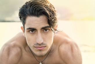 A young man with dark, wet hair looks directly at the camera with a serious expression. He is shirtless, showcasing a tanned and athletic build. The background is softly blurred, possibly depicting a beach or outdoor setting with bright, warm light illuminating the scene.