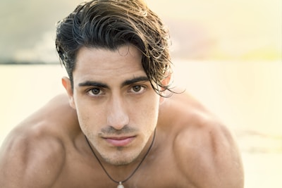 A young man with dark, wet hair looks directly at the camera with a serious expression. He is shirtless, showcasing a tanned and athletic build. The background is softly blurred, possibly depicting a beach or outdoor setting with bright, warm light illuminating the scene.