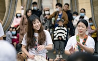 A group of people gathered at an outdoor event, with two women in the foreground playing traditional percussion instruments. The crowd in the background appears engaged, with individuals of various ages, some wearing masks.
