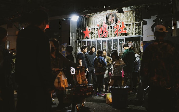 A group of people gather outside a storefront with glass doors, under signage with prominent red characters. The setting appears to be nighttime, and the storefront has bright lights illuminating the surrounding area. People seem engaged in conversation and activities, creating a lively atmosphere.