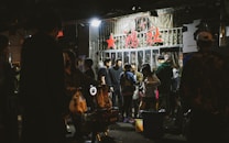 A group of people gather outside a storefront with glass doors, under signage with prominent red characters. The setting appears to be nighttime, and the storefront has bright lights illuminating the surrounding area. People seem engaged in conversation and activities, creating a lively atmosphere.
