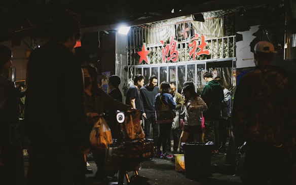 A group of people gather outside a storefront with glass doors, under signage with prominent red characters. The setting appears to be nighttime, and the storefront has bright lights illuminating the surrounding area. People seem engaged in conversation and activities, creating a lively atmosphere.