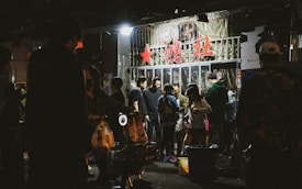 A group of people gather outside a storefront with glass doors, under signage with prominent red characters. The setting appears to be nighttime, and the storefront has bright lights illuminating the surrounding area. People seem engaged in conversation and activities, creating a lively atmosphere.