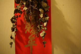 Dried leaves and twigs are hanging against a red background with Chinese characters. The composition includes earthy tones and creates a rustic, natural feel.