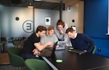 A group of four people gathered around a table with laptops, engaging in a collaborative and lively discussion. They are smiling and appear to be focused on something on the laptop screen. The setting is a modern office space with glass partitions and a relaxed atmosphere.