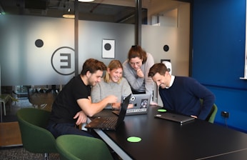 A group of four people gathered around a table with laptops, engaging in a collaborative and lively discussion. They are smiling and appear to be focused on something on the laptop screen. The setting is a modern office space with glass partitions and a relaxed atmosphere.