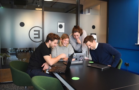 A group of four people gathered around a table with laptops, engaging in a collaborative and lively discussion. They are smiling and appear to be focused on something on the laptop screen. The setting is a modern office space with glass partitions and a relaxed atmosphere.