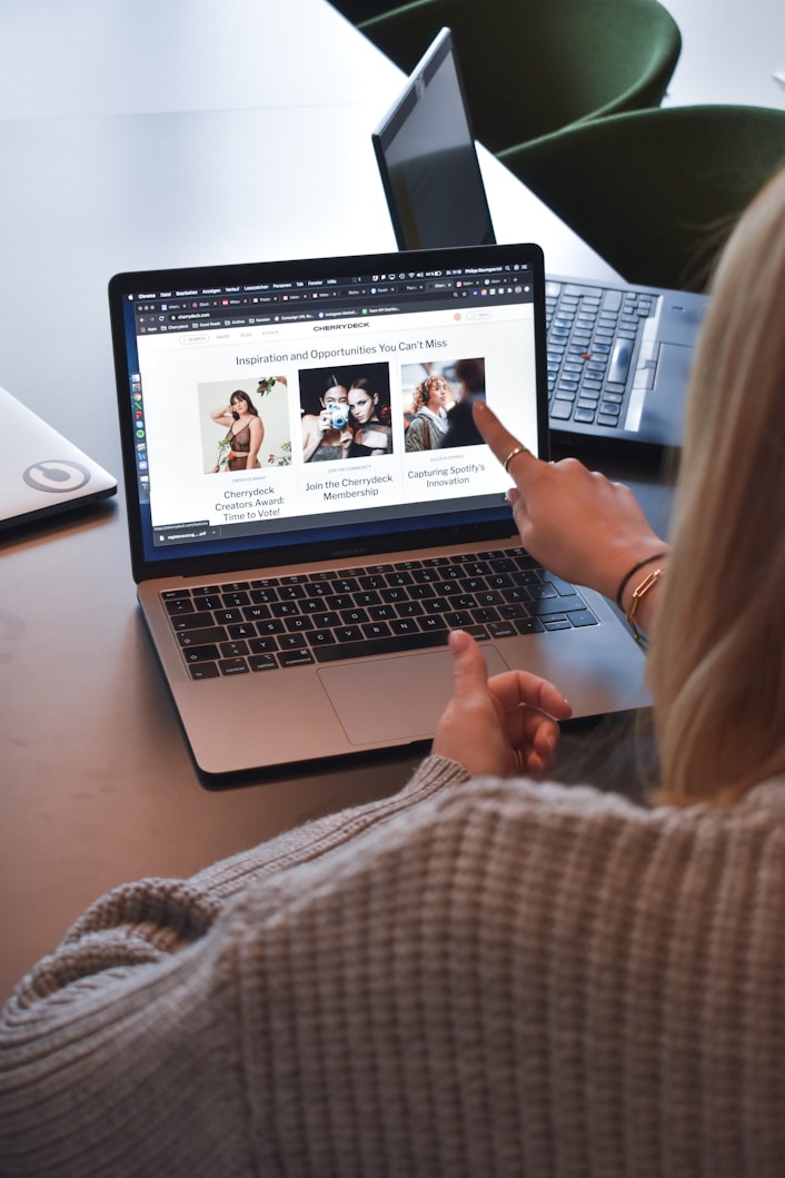 A person wearing a gray sweater is pointing to a laptop screen displaying a web page. The webpage contains a header titled 'Inspiration and Opportunities You Can't Miss' and features three sections with images and text. Nearby, there is another laptop on the table.
