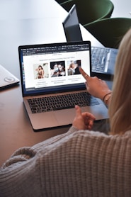 A person wearing a gray sweater is pointing to a laptop screen displaying a web page. The webpage contains a header titled 'Inspiration and Opportunities You Can't Miss' and features three sections with images and text. Nearby, there is another laptop on the table.