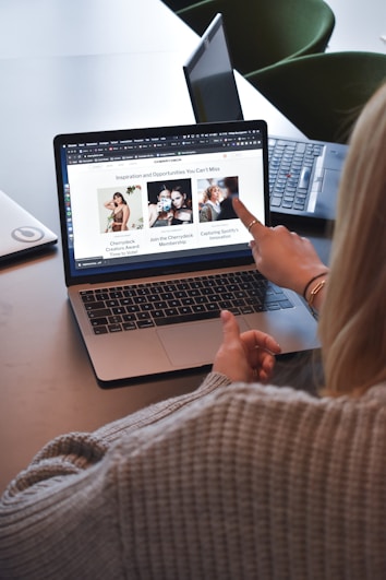 A person wearing a gray sweater is pointing to a laptop screen displaying a web page. The webpage contains a header titled 'Inspiration and Opportunities You Can't Miss' and features three sections with images and text. Nearby, there is another laptop on the table.