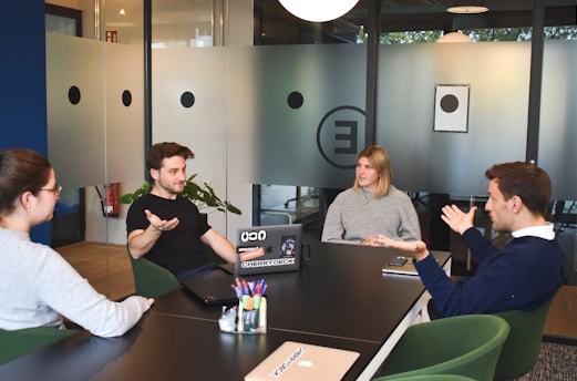 Four people are engaged in a discussion around a black conference table in a modern office setting. The room is enclosed with frosted glass walls that have circular designs. A laptop with several stickers is open on the table, and pens are organized in a holder. The individuals, two men and two women, seem to be engaged in a professional meeting or brainstorming session.