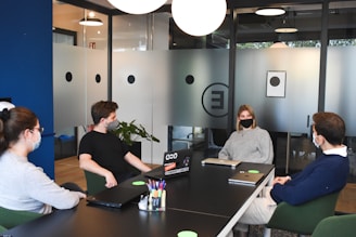 Four people wearing masks are seated around a conference table in a modern office setting. The room is softly lit, with glass walls featuring circular patterns. Laptops and notebooks are placed on the table, and a pen holder filled with various colored pens is visible. The atmosphere seems collaborative.