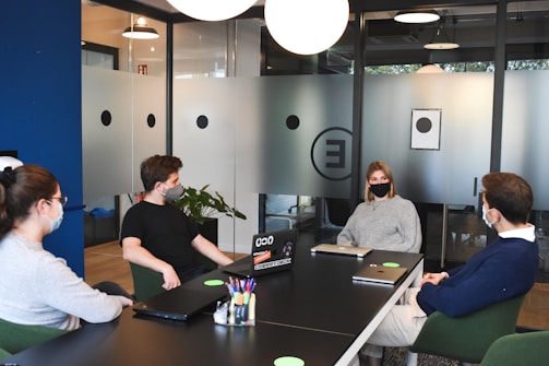 Four people wearing masks are seated around a conference table in a modern office setting. The room is softly lit, with glass walls featuring circular patterns. Laptops and notebooks are placed on the table, and a pen holder filled with various colored pens is visible. The atmosphere seems collaborative.