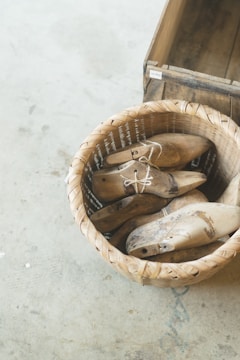 A wicker basket containing several wooden shoe molds is placed on a light concrete floor. Next to the basket, there is a wooden crate. The shoe molds are worn and have a rustic appearance, with some visible dirt and wear marks.