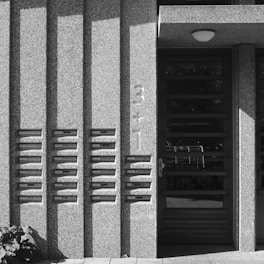 A concrete building facade features a row of mailboxes embedded in the wall, with shadows cast from the architectural elements. To the right is a modern door with a numeric address in a bold font reading '3+1'.