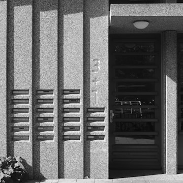 A concrete building facade features a row of mailboxes embedded in the wall, with shadows cast from the architectural elements. To the right is a modern door with a numeric address in a bold font reading '3+1'.
