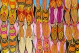 A vibrant array of traditional shoes displayed in an organized grid, featuring intricate embroidery and diverse patterns. The footwear exhibits an array of colors, including vivid pinks, oranges, yellows, and hints of blue, arranged on a flat surface, highlighting the craft and cultural richness.