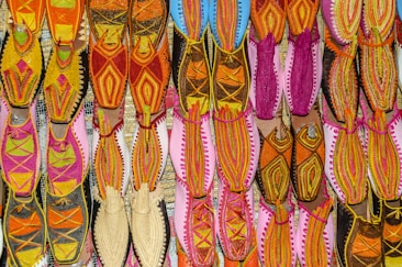 A vibrant array of traditional shoes displayed in an organized grid, featuring intricate embroidery and diverse patterns. The footwear exhibits an array of colors, including vivid pinks, oranges, yellows, and hints of blue, arranged on a flat surface, highlighting the craft and cultural richness.