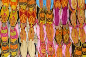 A vibrant array of traditional shoes displayed in an organized grid, featuring intricate embroidery and diverse patterns. The footwear exhibits an array of colors, including vivid pinks, oranges, yellows, and hints of blue, arranged on a flat surface, highlighting the craft and cultural richness.