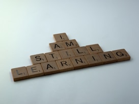 Wooden Scrabble tiles arranged to form the phrase 'I AM STILL LEARNING' against a plain background. The tiles are aligned in a triangular shape with the words intersecting each other.