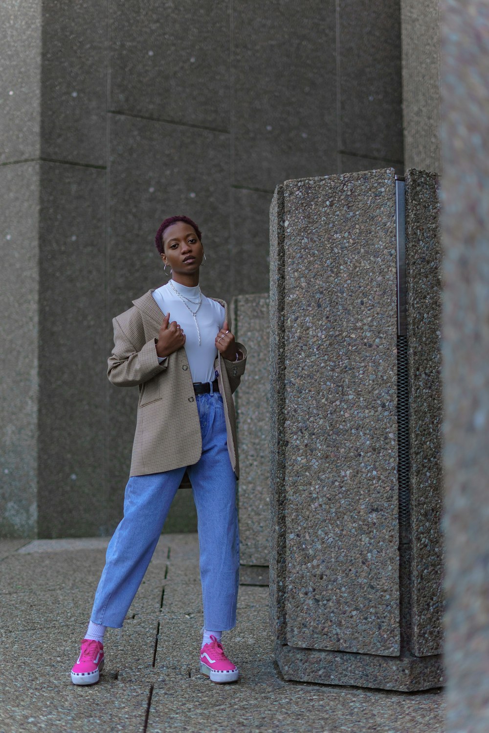 man in gray formal coat and blue denim jeans standing beside gray concrete wall