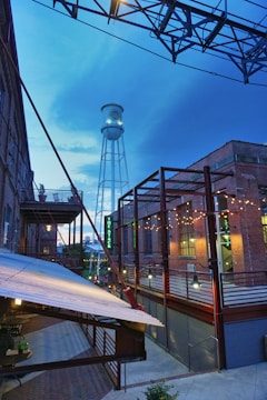 An industrial urban scene featuring a tall water tower against a blue sky, surrounded by brick buildings and iron frameworks. Strings of lights add warmth to the scene, while a staircase and a covered walkway can be seen in the foreground.