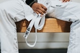 A person dressed in a white martial arts uniform is sitting on a wooden bench holding a white belt. The person's hands have some fingers wrapped with athletic tape.