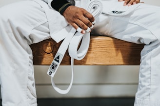 A person dressed in a white martial arts uniform is sitting on a wooden bench holding a white belt. The person's hands have some fingers wrapped with athletic tape.