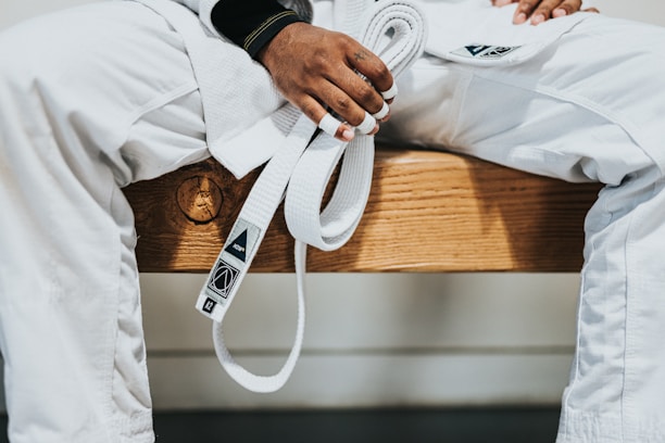 A person dressed in a white martial arts uniform is sitting on a wooden bench holding a white belt. The person's hands have some fingers wrapped with athletic tape.