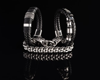 Two intricately designed bracelets composed of black braided leather and shiny metallic components are displayed against a dark reflective surface. One bracelet features a bold chain design with a detailed clasp resembling a wolf’s head.