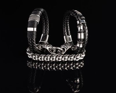 Two intricately designed bracelets composed of black braided leather and shiny metallic components are displayed against a dark reflective surface. One bracelet features a bold chain design with a detailed clasp resembling a wolf’s head.