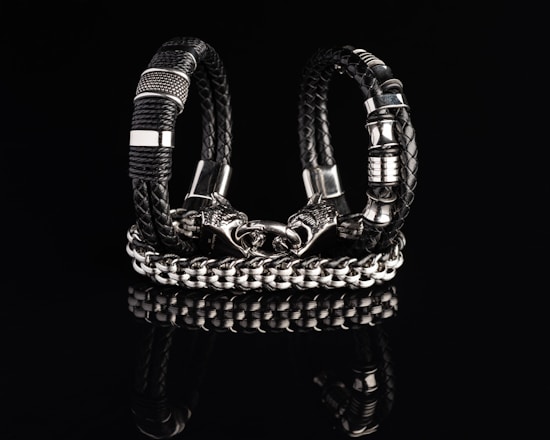 Two intricately designed bracelets composed of black braided leather and shiny metallic components are displayed against a dark reflective surface. One bracelet features a bold chain design with a detailed clasp resembling a wolf’s head.